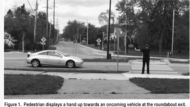 Figure 1.  Pedestrian displays a hand up towards an oncoming vehicle at the roundabout exit.  Picture shows man standing at the edge of the crosswalk, wearing black pants, shirt and baseball cap, and holding a white cane with the tip close to his left foot.  He is facing forward and holding his left arm horizontal, with his hand up and palm facing the car that is approaching from his left.